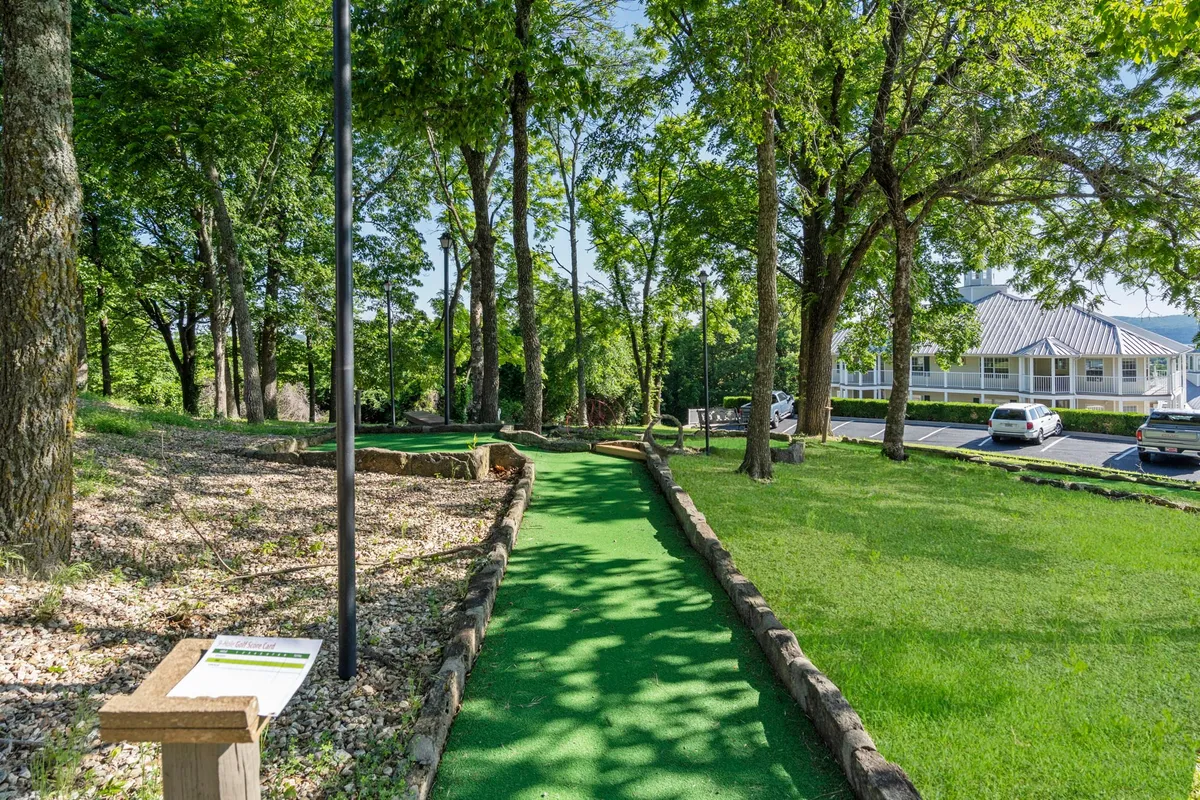 A cheerful exterior view of the resort's mini-golf course, framed by lush trees and dotted with families enjoying a game. The image captures the inviting greens, the informative sign, and hints of parked cars in the background, creating a scene full of activity and playfulness.