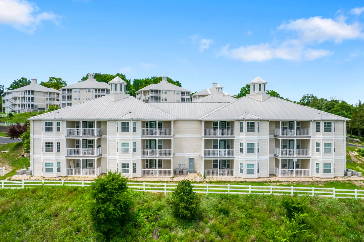 This exterior image features the building structure with a beautifully landscaped area and a white fence. The scene includes people relaxing on balconies and enjoying the open green space, with the backdrop of a peaceful, wooded environment.