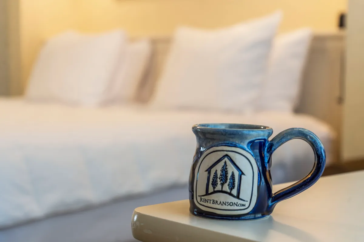 A cozy king bedroom setting featuring a neatly made bed with white linens and a ceramic mug on the side, suggesting a relaxing morning routine. The room exudes warmth and tranquility, making it an ideal place to unwind after a busy day by the lake or pool.