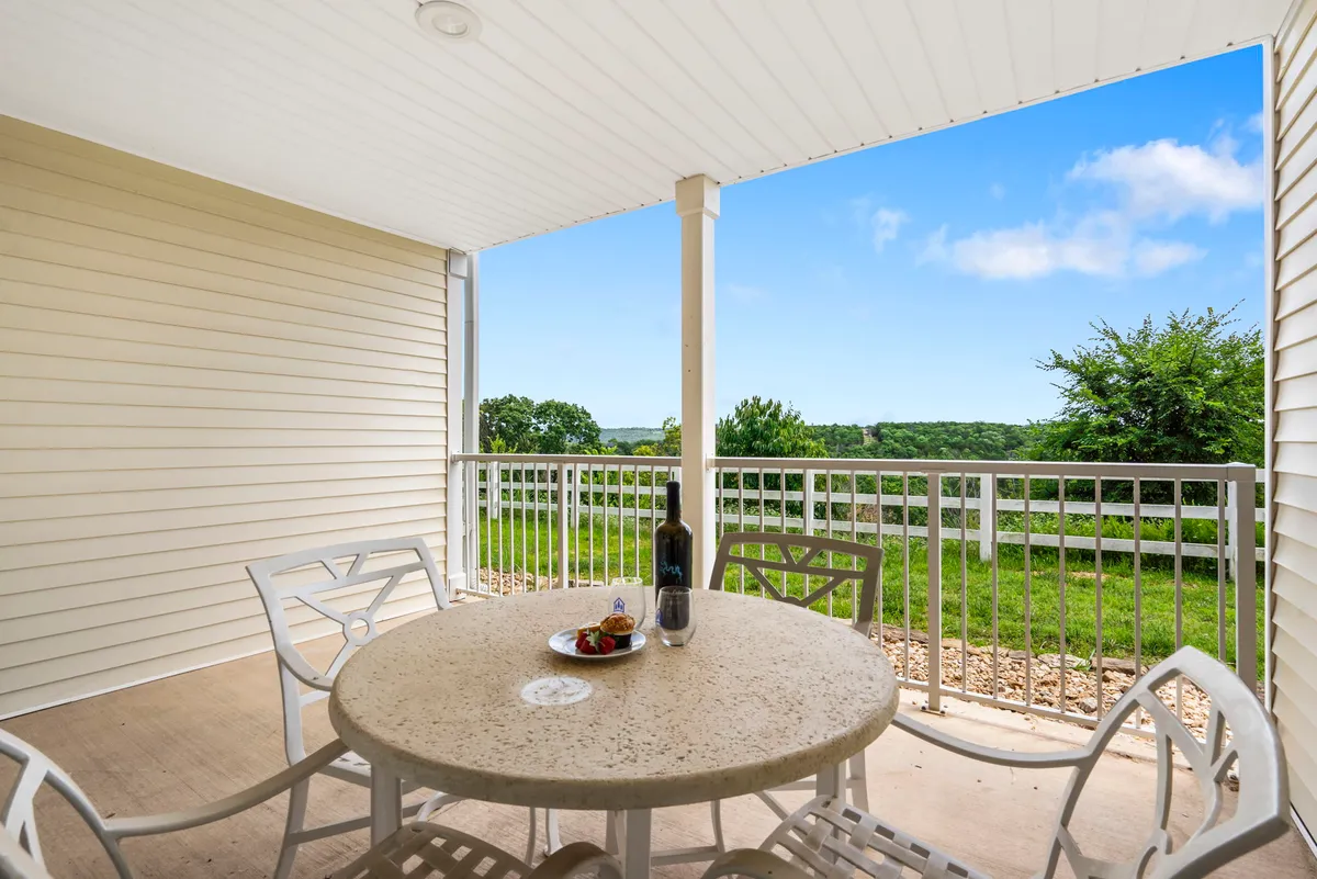 A cozy deck scene with a table set with fruit and wine, overlooking a scenic view framed by a fence and lush greenery. The setting glows with soft natural light, hinting at either sunrise or sunset, and feels both inviting and tranquil—perfect for morning coffee or evening relaxation.