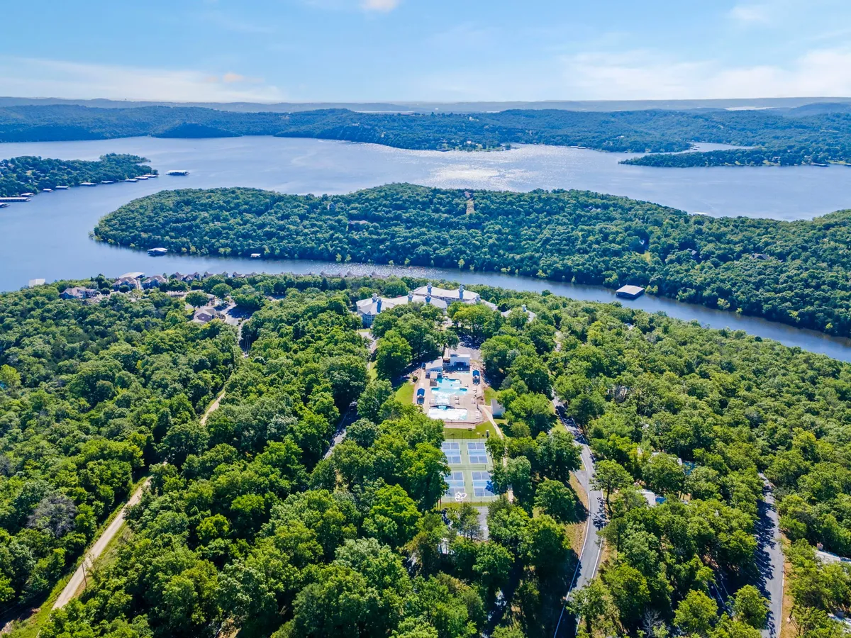 A wide exterior shot featuring lush green trees, sparkling water, a swimming pool, tennis courts, and residential buildings in the background. The scene is tranquil, with the soft colors of either sunrise or sunset reflected on the water, reinforcing a lakeside retreat atmosphere.