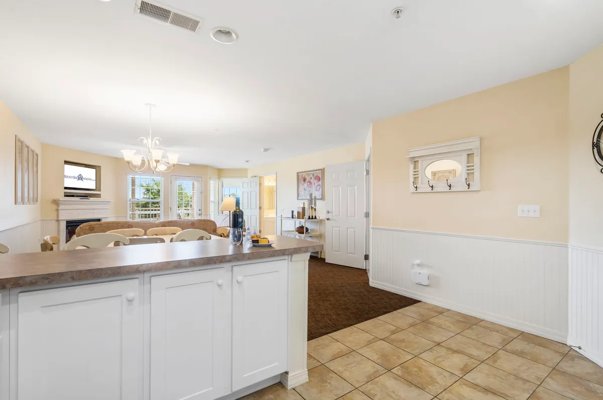 This bright interior image showcases an open-concept space seamlessly connecting the kitchen island, dining area, and cozy living area with a visible fireplace. Sunlight streams in through the windows, highlighting the airy and welcoming atmosphere. A doorway leads outside, reinforcing the home's open, interconnected feel.
