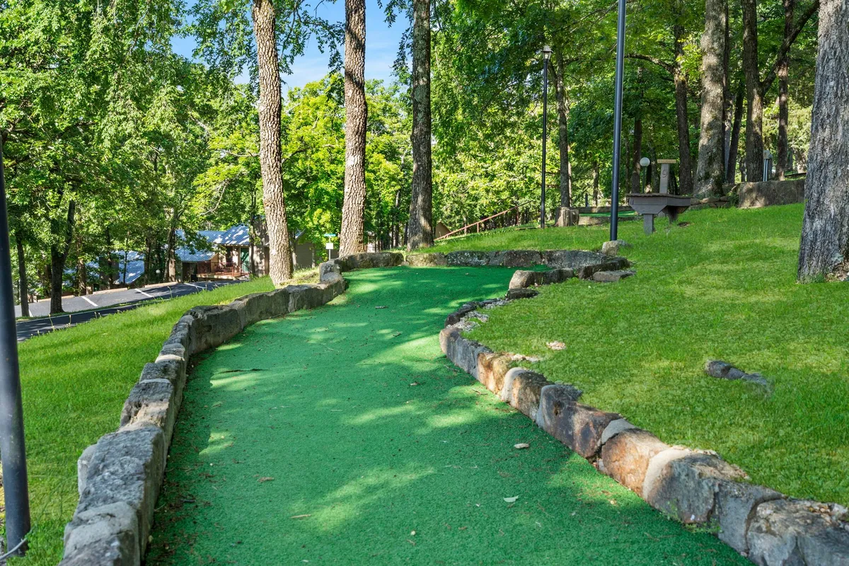 A vibrant mini-golf course is nestled among mature trees, with stone borders outlining the green pathways. The area feels playful and inviting, perfect for families or groups enjoying a friendly game amidst lush surroundings.
